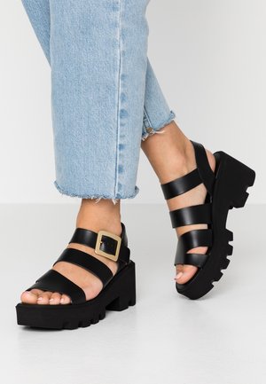 Sorte platformer sandaler med en chunky sål, der har flere stropper og en firkantet guldbuckle. Materialet ser glat og mat ud.