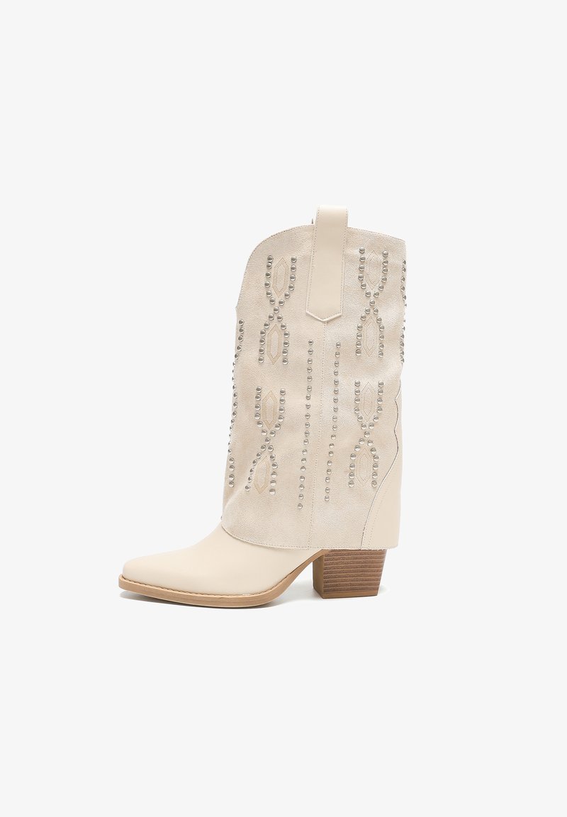 Botas de vaquero beige con punta afilada, parte superior de ante, tachuelas decorativas plateadas en un patrón de hoja y un tacón de madera apilada.