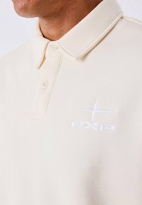 Lichtbeige polo-shirt van zachte stof, met een klassieke kraag en twee knopen. Op de borst is een wit geborduurd logo weergegeven.