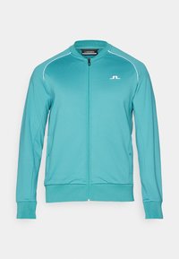 Teal zip-up jakna z ribastim ovratnikom in manšetami, bele akcentne črte vzdolž ramenskih delov, z stranskimi žepi in logotipom na prsih.