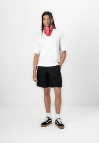 T-shirt blanc à manches courtes, short noir, bandana rouge et baskets noires avec des accents blancs. Le modèle se tient devant un fond uni.
