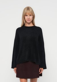 Pull noir en maille texturée, à la coupe ample, avec des épaules tombantes et un col rond ; porté par-dessus une jupe bordeaux. Matière douce et duveteuse.