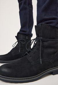 Botas de ante negro con cordones y cremallera lateral, con punta redondeada y superficie texturizada, sobre unos jeans de denim oscuro.
