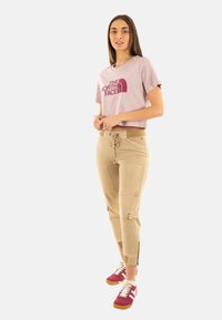 T-shirt rosa corto con logo bordeaux, pantaloni beige con tasche laterali e zip, abbinati a sneaker rosse e bianche con suola di gomma.