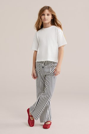 Jeune fille aux longs cheveux portant un haut blanc à manches courtes, un pantalon rayé noir et blanc et des chaussures rouges, debout les jambes croisées.