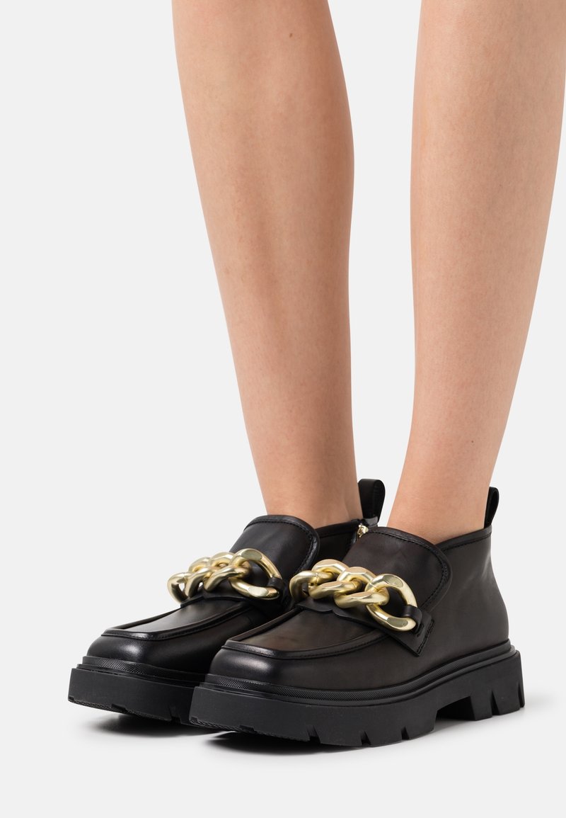 Mocassins noirs à semelle épaisse avec un détail de grosse chaîne dorée, portés sur des jambes nues sur fond blanc.