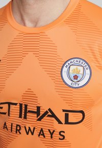 Maglia da portiere arancione con un design testurizzato. Presenta testo nero per "ETIHAD AIRWAYS" e un logo circolare blu del Manchester City.