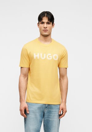 Man wearing a yellow T-shirt with "HUGO" text, paired with light blue jeans, standing against a plain gray background.