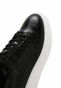 Sneaker in pelle nera con texture di coccodrillo, punta rotonda e suola in gomma bianca. Presenta lacci neri e dettagli cuciti lungo i lati.