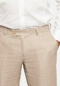 Pantalones de lino beige con un sutil patrón texturizado, que cuentan con un cierre de botón en la parte frontal y bolsillos laterales. Corte ajustado, diseño de tiro medio.
