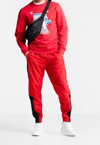 Personne portant un sweat-shirt rouge NBA All-Star, un pantalon de survêtement rouge et noir, des baskets blanches et un sac bandoulière noir, debout les mains dans les poches.