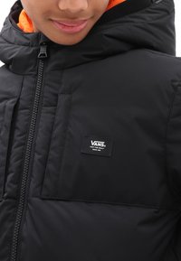 Persona que lleva una chaqueta con capucha negra de Vans con un forro naranja y un cierre en la parte delantera, mostrando el parche del logo de Vans en el pecho.