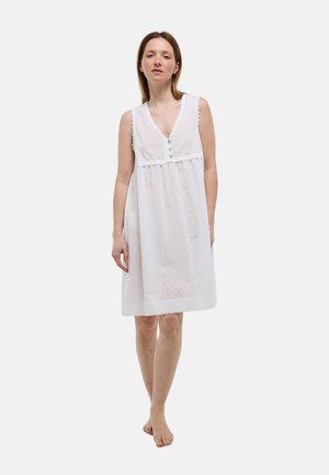 Femme debout pieds nus portant une robe blanche sans manches avec des boutons et un motif texturé sur un fond clair uni.