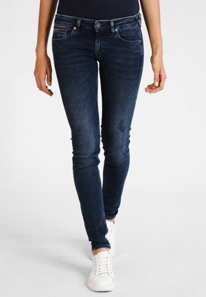 Jeans skinny de color azul oscuro con un ligero desteñido, bolsillos frontales y cierre de botón, combinados con zapatillas blancas. Textura de denim suave.