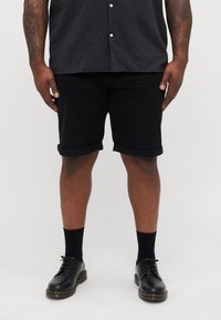 Zwarte katoenen shorts met opgerolde boorden, gecombineerd met zwarte veterschoenen en enkelsokken, gedragen door een persoon in een donker shirt.