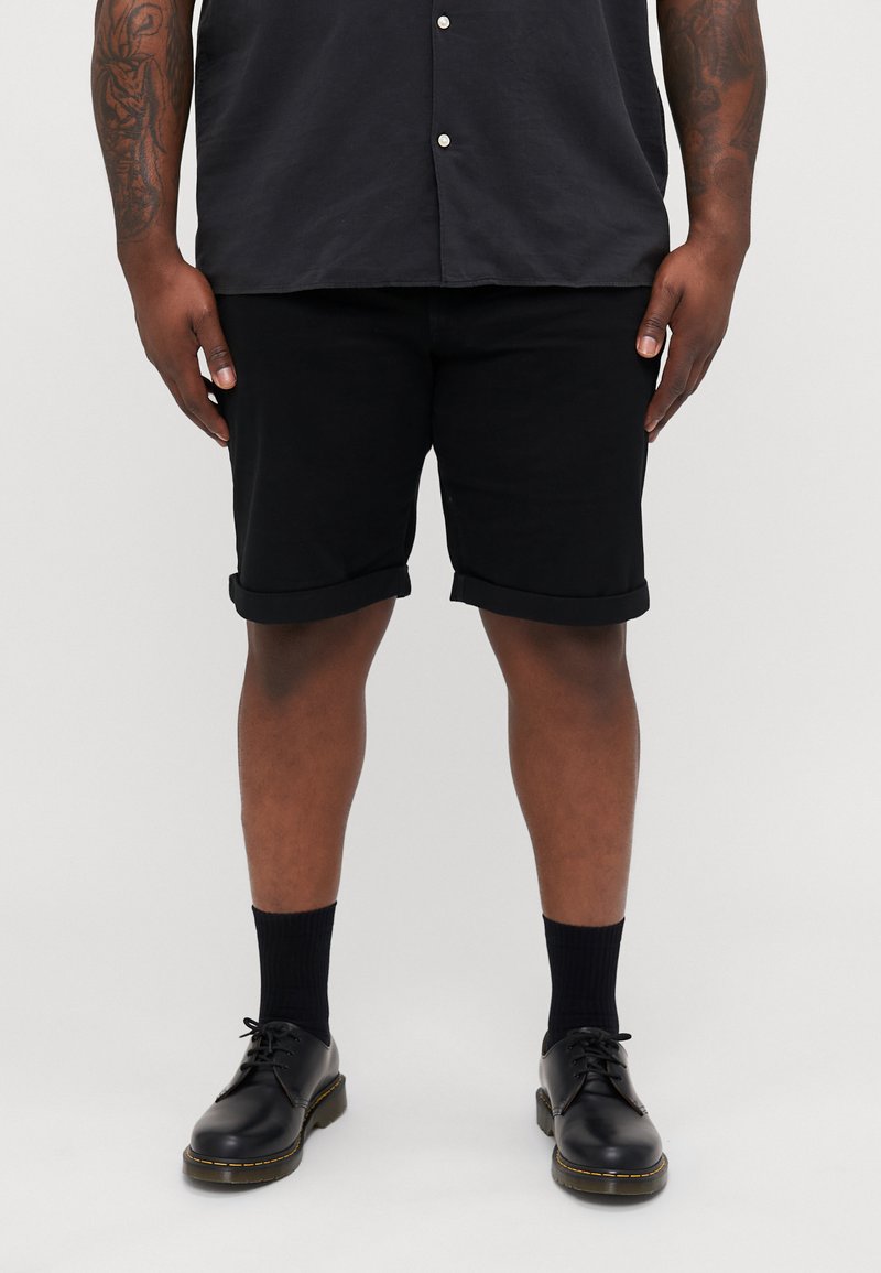 Zwarte katoenen shorts met opgerolde boorden, gecombineerd met zwarte veterschoenen en enkelsokken, gedragen door een persoon in een donker shirt.