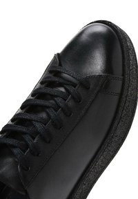 Black leather sneaker featuring a smooth upper, round toe, textured black rubber sole, and black flat laces. Minimalist design with subtle stitching.