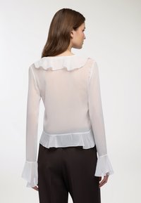 Camicia bianca trasparente con collo e polsini arricciati, maniche lunghe e orlo svasato. Tessuto leggero e strutturato con pieghe sottili.