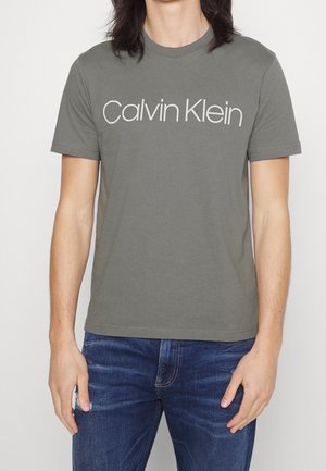 Homme portant un T-shirt gris à manches courtes avec logo Calvin Klein blanc et un jean bleu, debout les bras détendus.