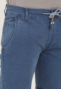 Sinine denim püksid tekstureeritud viimistlusega, küljetaskudega ja kontrastset värvi paelaga õmmeldud vöökoht, mis võimaldab reguleerimist.