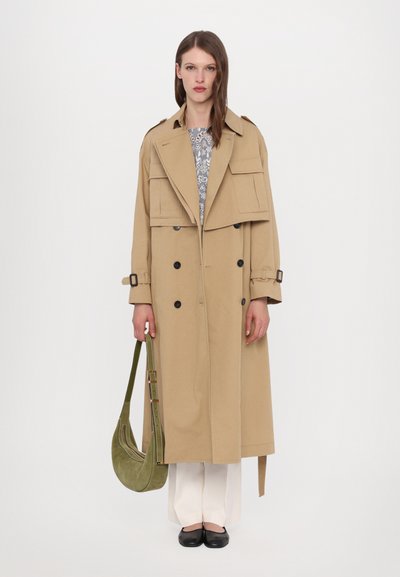 Trench coat beige con design doppiopetto, grandi tasche e colletto ampio. Abbinato a pantaloni bianchi e scarpe nere flat, mentre tiene una borsa verde.
