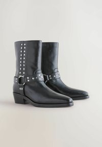 Botas negras de cuero hasta el tobillo con punteras afiladas, acentos de cinturón plateado con tachuelas y un tacón bajo. Textura suave con tachuelas decorativas en el lateral.
