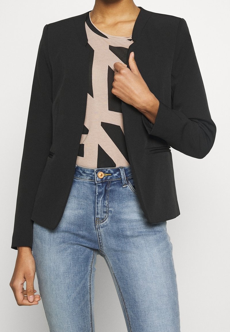 Personne portant un blazer noir ouvert sur un haut beige à motifs avec un jean bleu, tenant le revers du blazer d'une main.