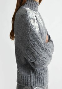 Grauer Strickpullover mit hohem Kragen, strukturiertem Muster und dekorativen Paillettenverzierungen an den Schultern. Weiches, lockeres Design.