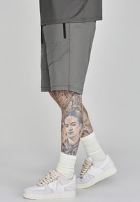 Grijze sportshorts met zijzakken en gestikte details, gecombineerd met witte sneakers en geribde witte sokken. Gedetailleerde tatoeages op het been.