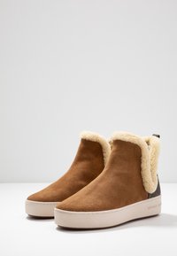 Bottines en daim marron avec un doublure en fausse fourrure crème, une semelle plate beige et un accent en tissu gris à l'arrière.