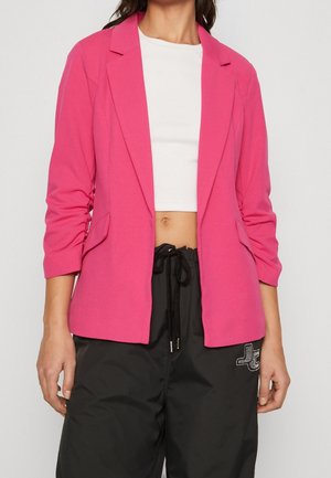 Vrouw draagt een felroze blazer over een wit cropped top, gecombineerd met losse zwarte broek met trekkoord en een klein logo op de rechterdij.