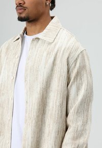 Lichtgekleurd, gestructureerd overhemd met verticale strepen, voorzien van een kraag en zichtbare knopen, gedragen over een wit shirt.