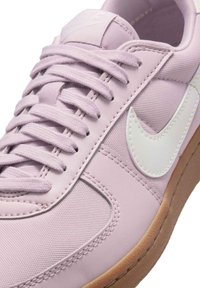 Ljuslila sneaker med texturerat tyg, vit swoosh och brun gummisula. Har platta snören och en vadderad tunga.