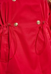 Rode stoffen jas met een vergrendelde taille, met gouden knoopaccenten en trekkoorddetails bij de taille en zakken. Gladde textuur.