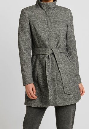 Femme portant un manteau en laine gris mi-cuisse avec un col haut, des manches longues, des poches latérales et une ceinture nouée à la taille.