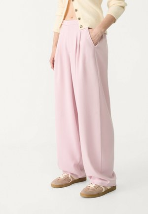 Persoon die lichtroze wijde broek, beige sneakers en een crèmekleurig vest met knopen draagt, met de hand in de zak.