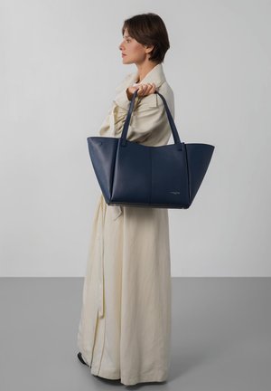 Femme portant un long trench-coat crème, tenant un grand sac cabas en cuir bleu marine sur l'épaule, debout devant un fond uni.