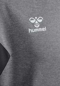 Szara tkanina o fakturze z białym logo "hummel" przedstawiającym stylizowaną pszczołę i czarny kształt jodełki po lewej stronie.