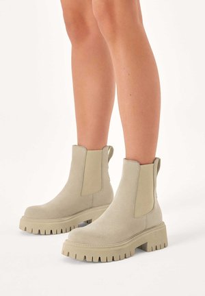 Bottes à cheville en suede gris clair avec des panneaux latéraux en élastique, semelle en caoutchouc épaisse et plate, et un détail texturé à rainures sur la semelle.