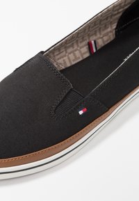 Scarpa nera in tela da indossare facilmente con rifiniture marroni, suola bianca con una striscia nera, e piccolo logo in tessuto rosso, bianco e blu sul lato.