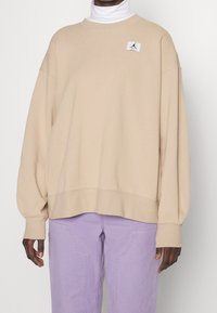 Sweat-shirt beige oversize à manches longues, col rond, et écusson logo. Associé à un pantalon violet clair avec poches latérales.