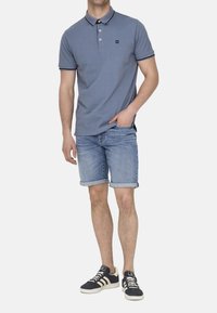 Blaue Poloshirt mit einem Kragen mit zwei Knöpfen, marinefarbigen Akzenten, kombiniert mit Jeansshorts. Modell trägt dunkle Sneakers mit weißen Akzenten.