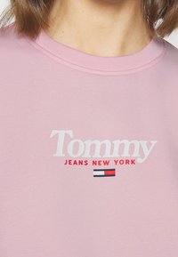 Vaaleanpunainen collegepaita, jossa on pyöreä kaula-aukko, resorikaulus ja valkoinen kirjailtu teksti "Tommy JEANS NEW YORK" punaisilla yksityiskohdilla sekä pieni lippulogo.