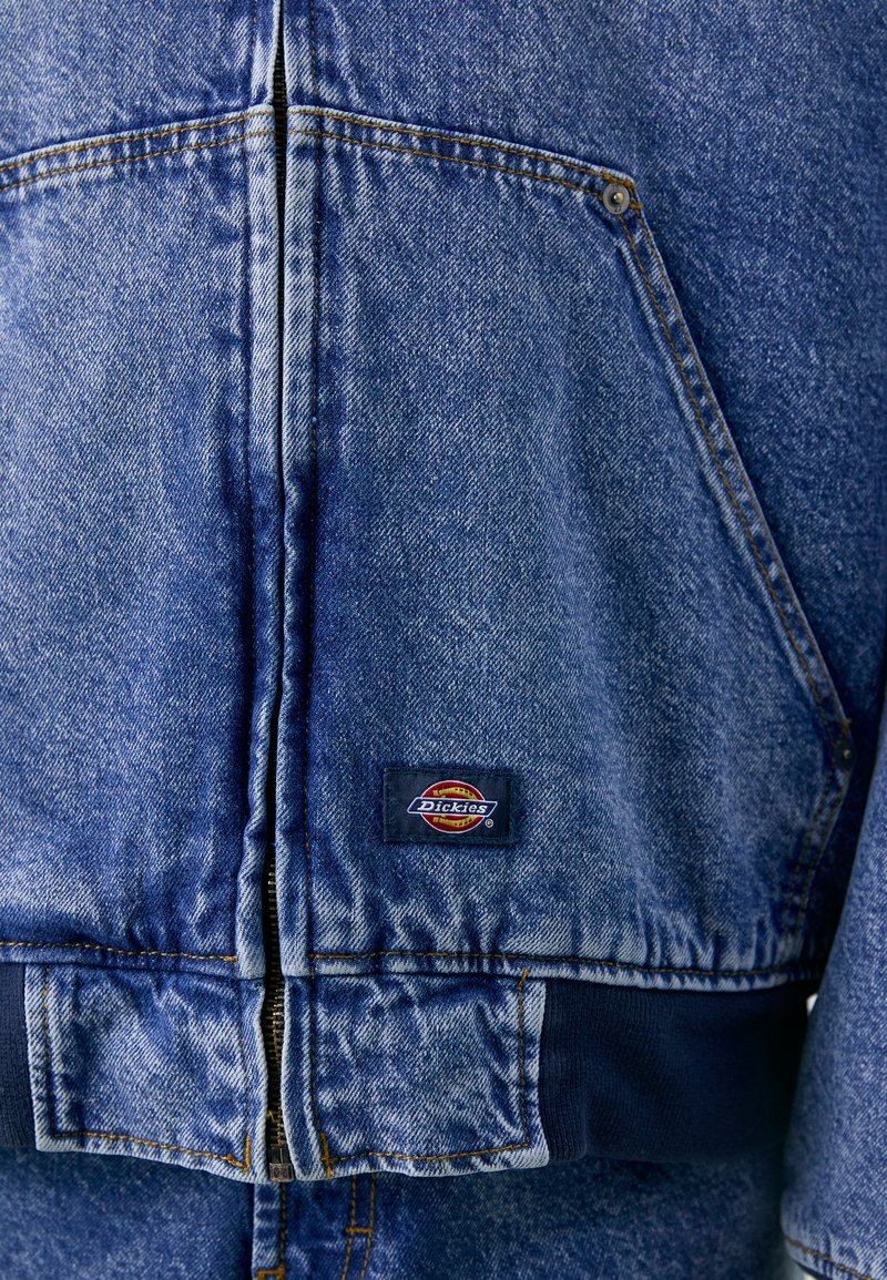 Giacca di jeans di colore azzurro chiaro con una texture sbiadita, dotata di chiusura a zip, vita a coste e una patch nera del logo Dickies sulla tasca sinistra.