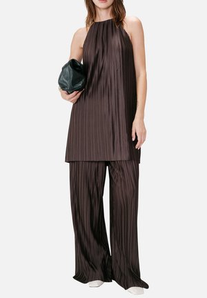 Kvinde iført ærmeløs mørkebrun plisseret tunika og matchende bukser med vide ben, der holder en sort clutch og står på hvid baggrund.