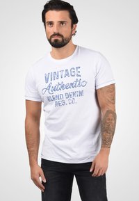 Bela bombažna majica s kratkimi rokavi, okroglim izrezom in sproščenim krojem, z napisom 'VINTAGE Authentic Blend Denim REG. CO.' v modrih črkah.