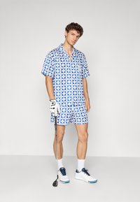 Kratek rokav modra srajca s vzorcem in ujemajoče se kratke hlače, z okroglimi motivi v modri, beli in rumeni barvi. Model drži golf palico.