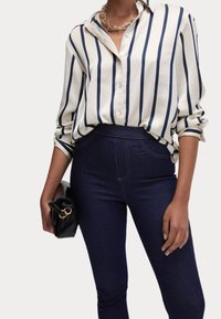 Camisa abotonada de color beige claro con rayas verticales azules marino, combinada con jeans de tiro alto de color azul oscuro. Sosteniendo un bolso negro con herrajes dorados.