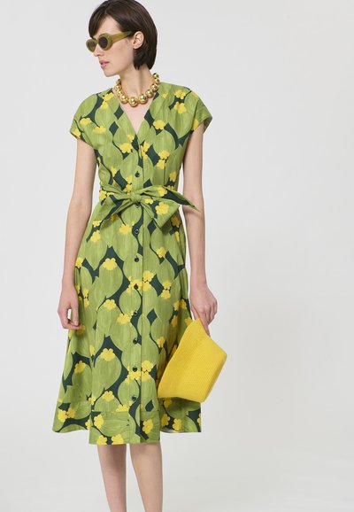 Femme portant une robe fleurie verte et jaune avec une taille nouée, des lunettes de soleil vertes, un collier de perles dorées, et tenant une pochette tissée jaune.
