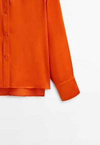 Chemise orange vif à manches longues boutonnée, avec un poignet pointu et un ourlet légèrement incurvé sur fond blanc.
